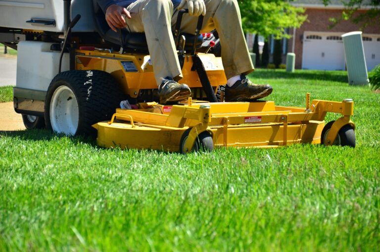 A person sits on a yellow riding lawn mower, providing expert lawn maintenance as they cut green grass in a residential yard. Their legs and feet are visible, along with spinning blades. A house and driveway appear in the background.