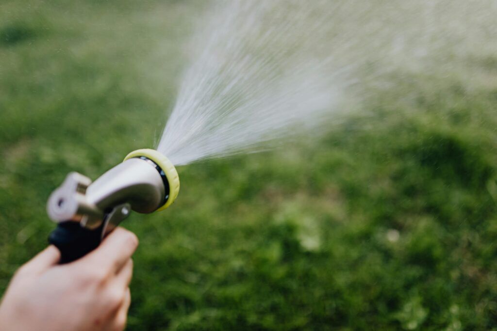 A hand holding a garden hose nozzle sprays water in a fine mist over a grassy lawn. The background is blurred, focusing on the watering action.