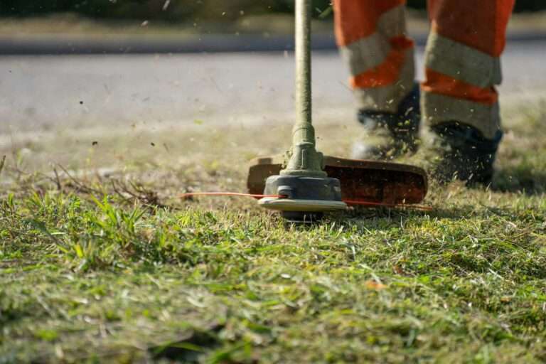 A person wearing orange safety pants uses a grass trimmer to cut grass on a lawn. Grass clippings and debris are visible in the air near the rotating trimmer head.